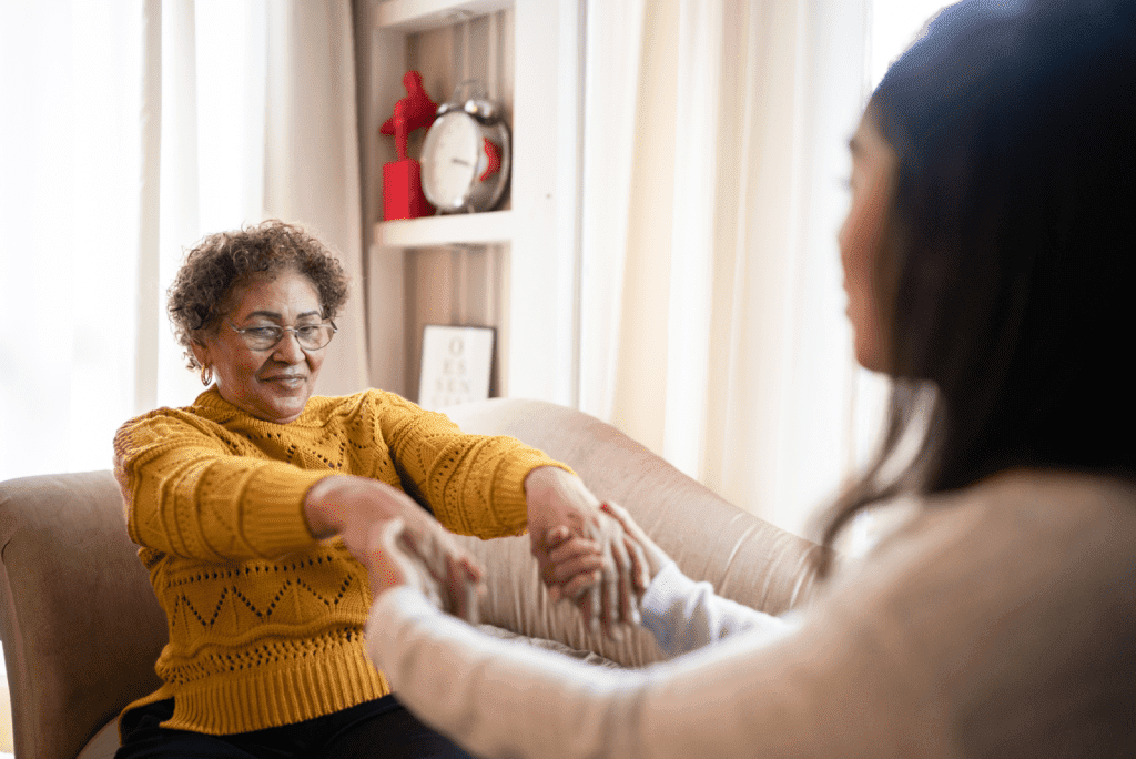 Caregiver helping senior woman in a mustard cardigan off the couch