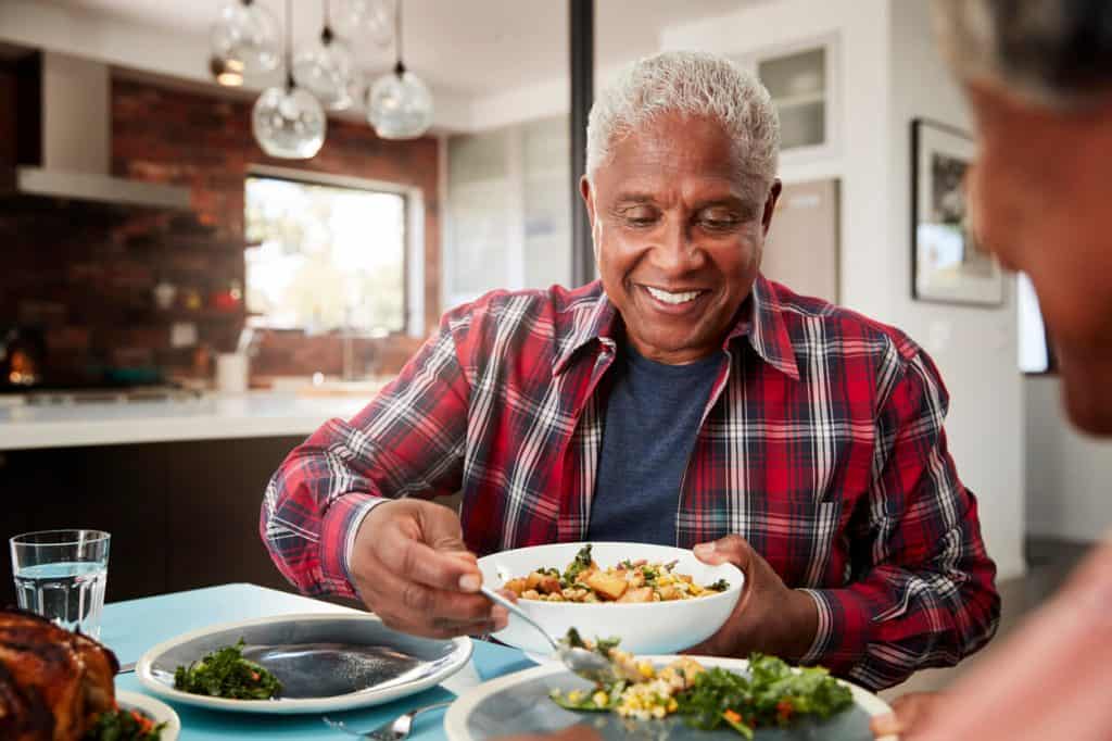 What is the difference between assisted living and nursing home. Senior man smiling and enjoying a delicious home cooked meal sitting at a table.