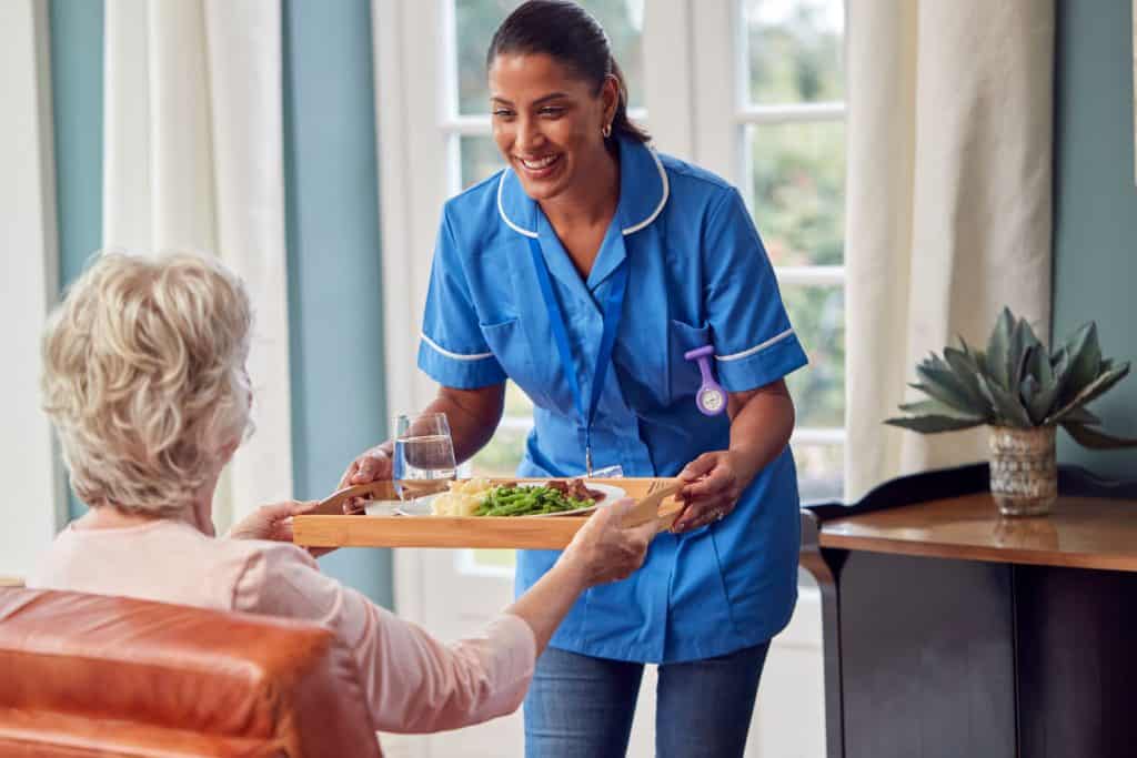 Female caregiver in retirement facility bringing a meal on a tray to a senior woman