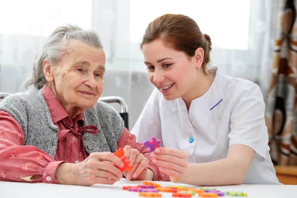 independant caregiver jobs: Young female caregiver playing jigsaw puzzle with an elderly woman at home