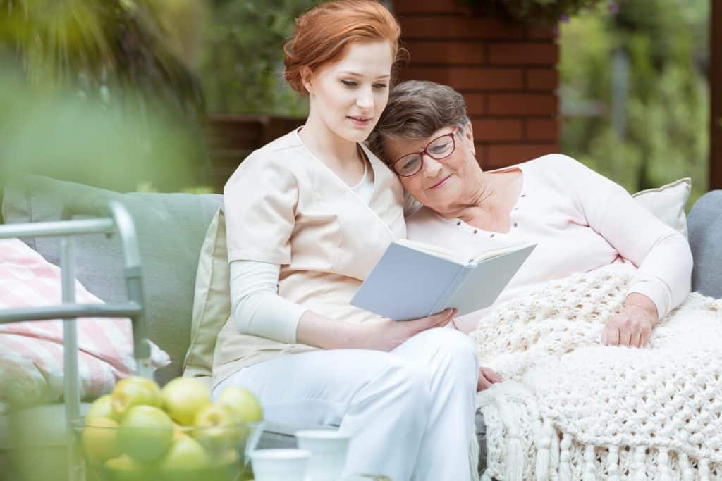 private live in caregiver jobs A live-in caregiver reading a book to a senior woman.