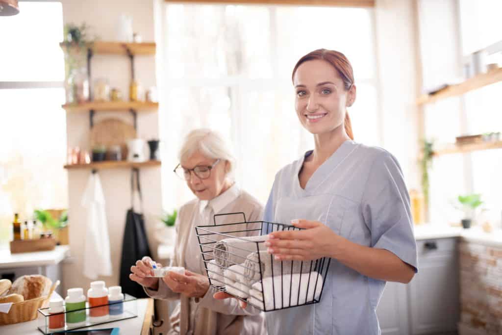 A live-in caregiver helping a senior woman organize her medications while holding a basket with folded towels.