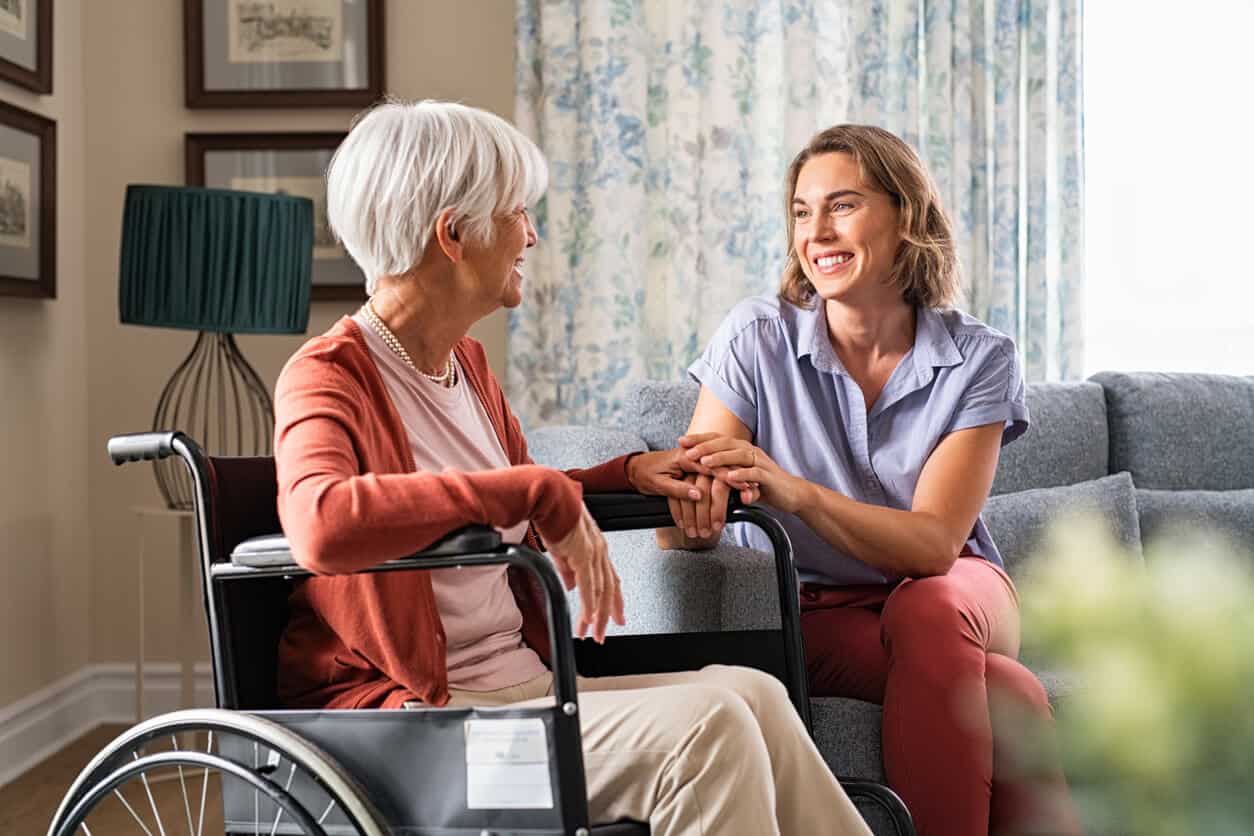 A live-in caregiver is smiling to a senior woman in a wheelchair while holding her hand. live in caregiver jobs near me