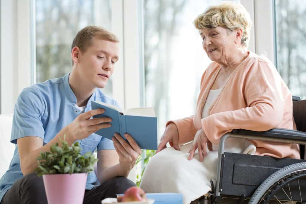 Male in-home caregiver reading a book to an elderly woman at home