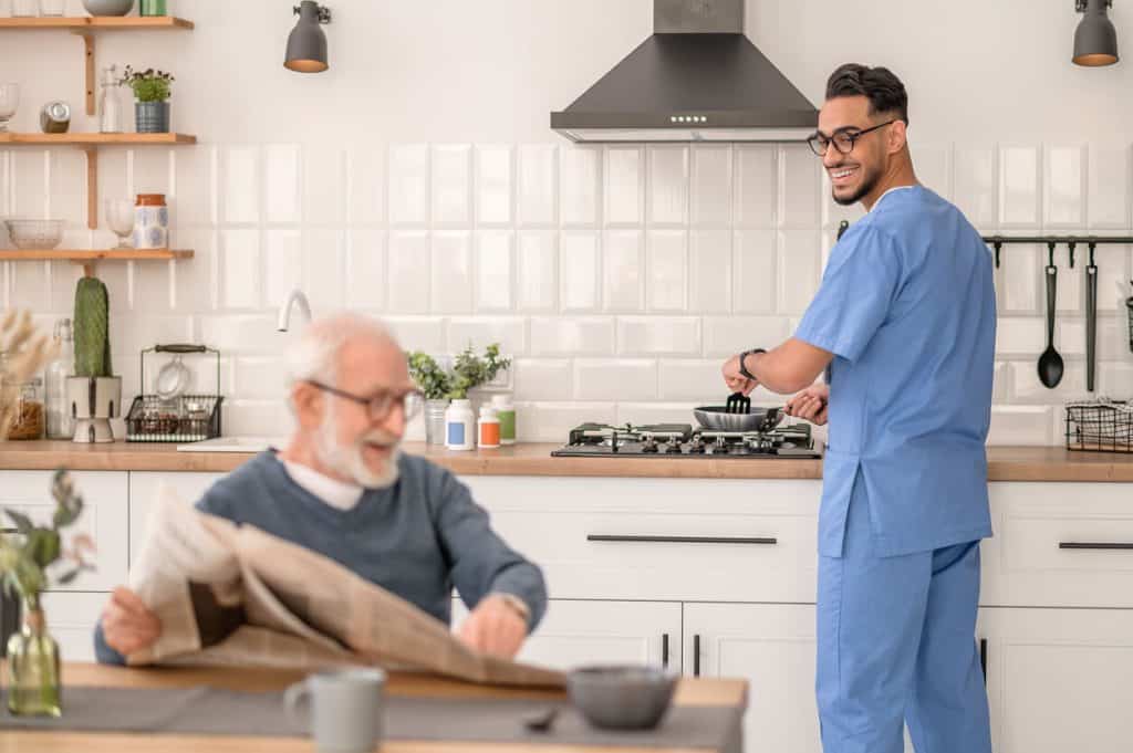 Male home care provider cooking while an elderly person is sitting at the dining table