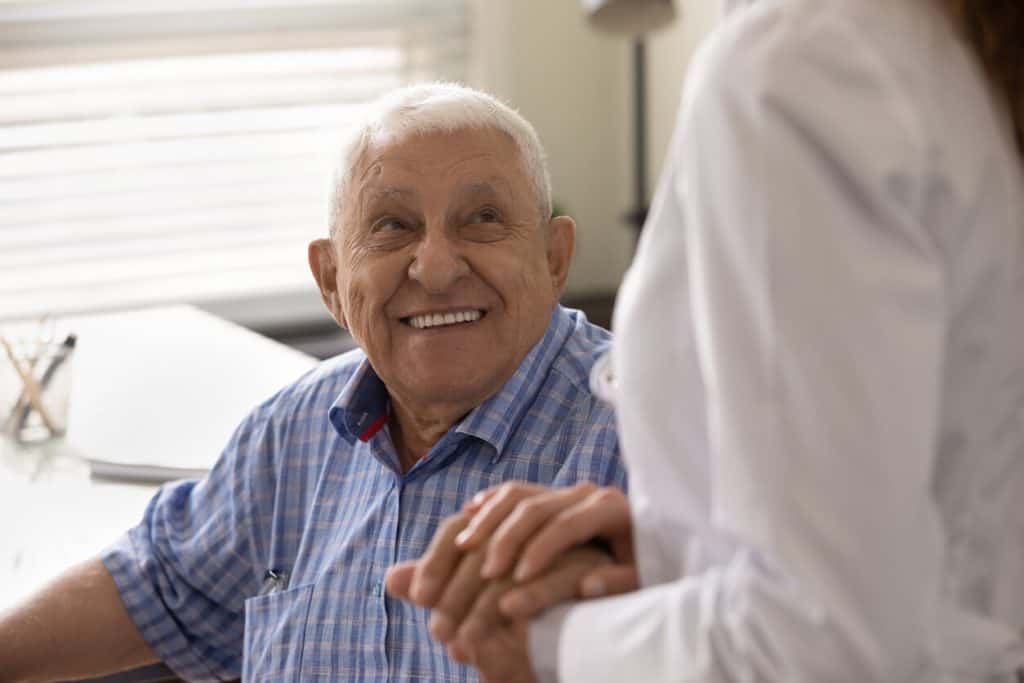 Elderly man smiling up and holding the hand of his caregiver, mental health assisted living facilities.