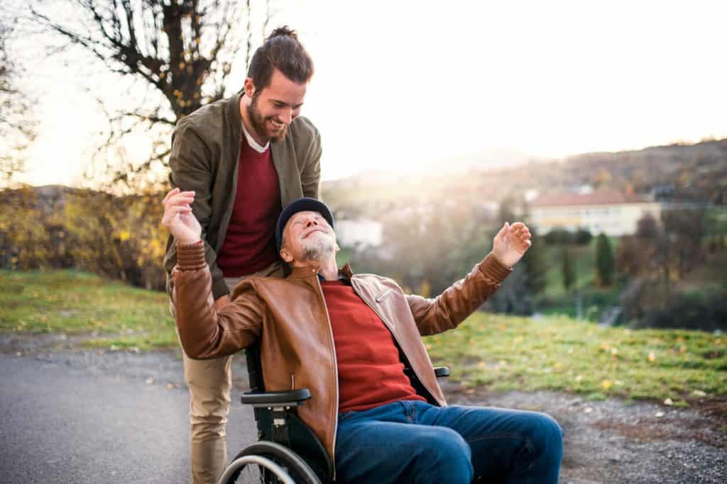 Young male caregiver talking to joyful senior man as caregiver pushes him in wheelchair, jobs taking care of elderly