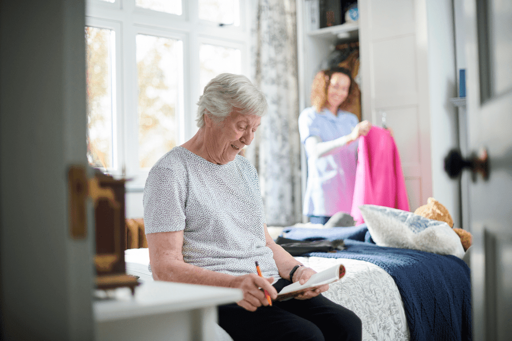 An elderly woman is looking at her notebook while female caregiver is organizing her closet in the background 2022, home care nursing