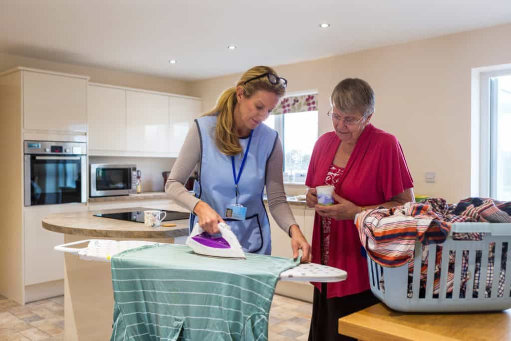 Homecare assistance helping an elderly woman to iron clothes at home 2022, assistance home care