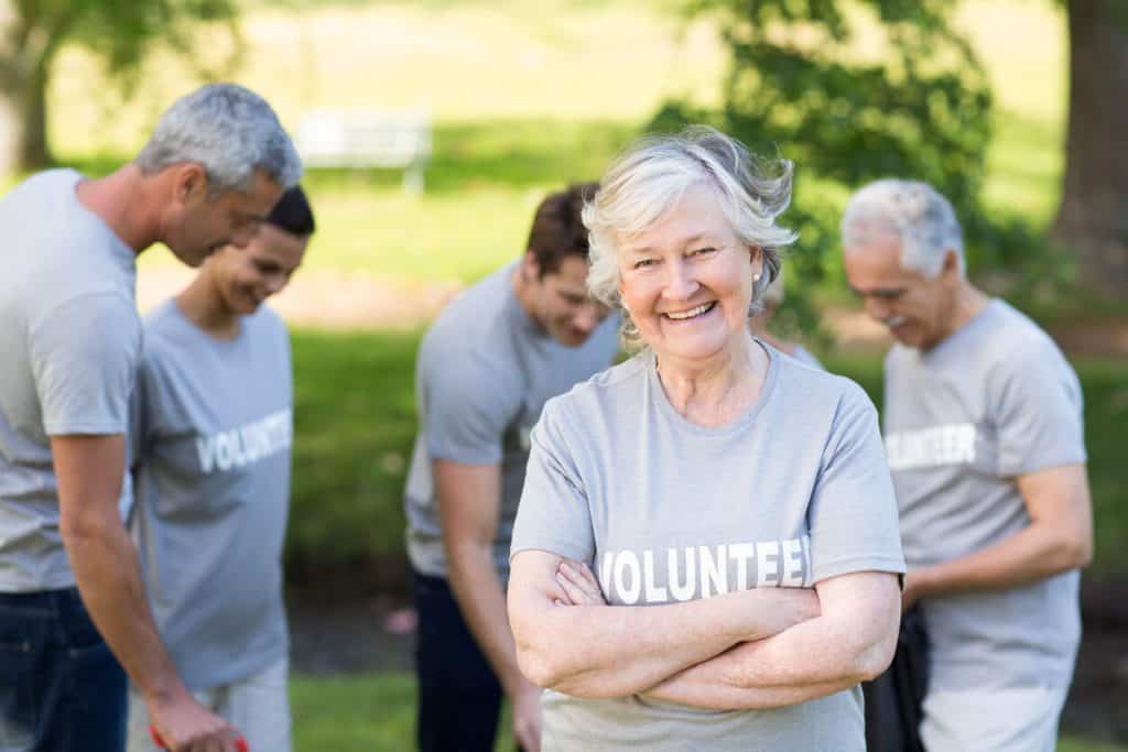 A senior woman smiling at the camera with her group of other volunteers cleaning the park. unique activities for seniors