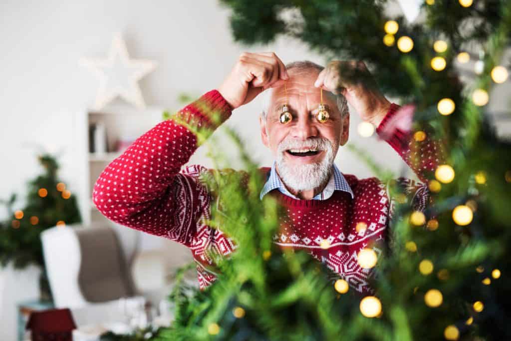 Senior man dressed for Christmas standing by a Christmas tree and holding two ornaments in front of his eyes 2022 christmas activities for seniors