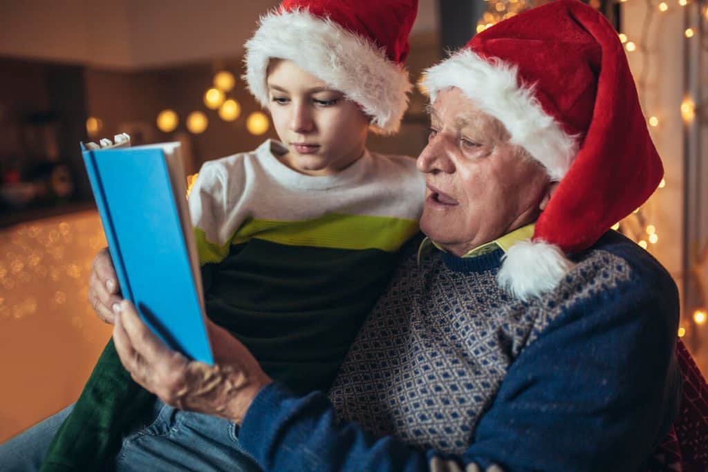 Grandfather reading the Christmas story to his grandson in Santa hats. Christmas activities for seniors