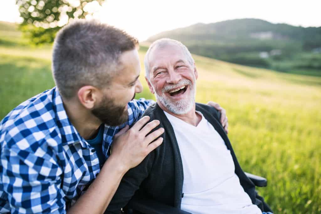 Adult male child pushing dad in wheelchair outside and they are laughing memory care activities for seniors
