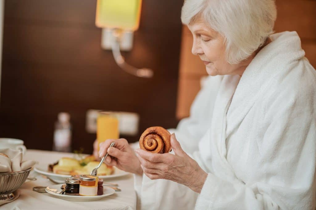 Senior woman in white robe eating delicious breakfast food, luxurious assisted living