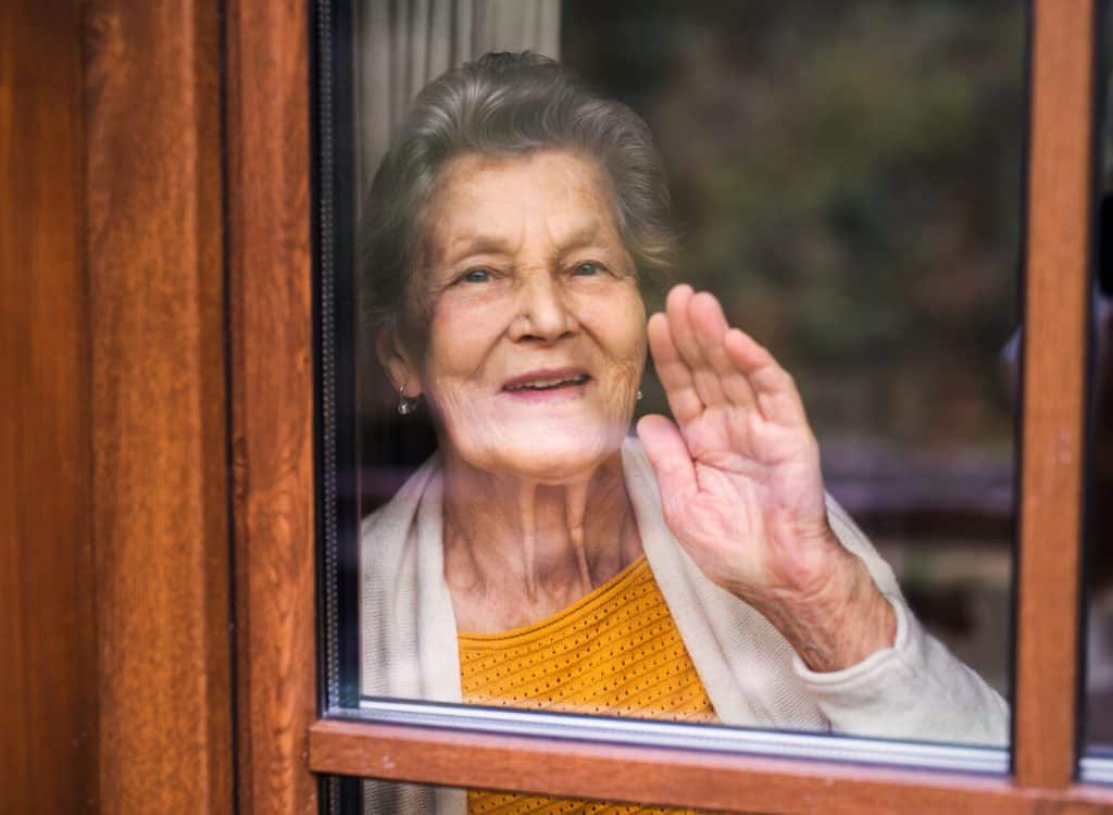 A happy elderly woman standing by the window, looking out and waving through the glass.