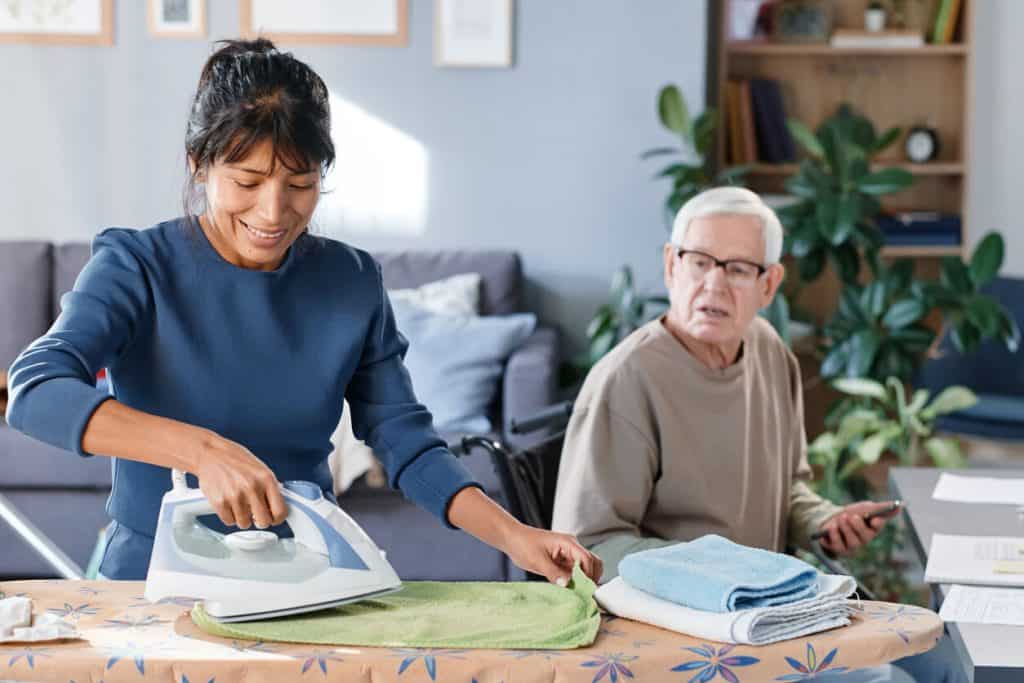 Smiling caregiver providing support and talking to senior man, she is helping him with household chores, home assisted living