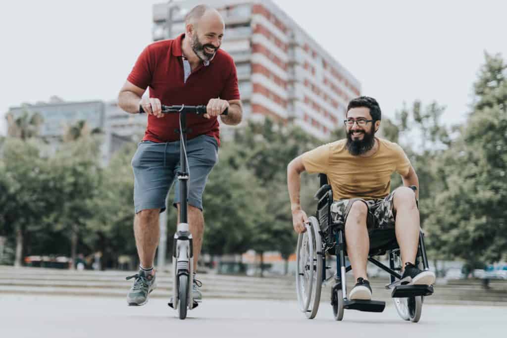 Caregiver riding a scooter alongside disabled person in a wheelchair. Both are smiling 2022, inhome care services for disabled