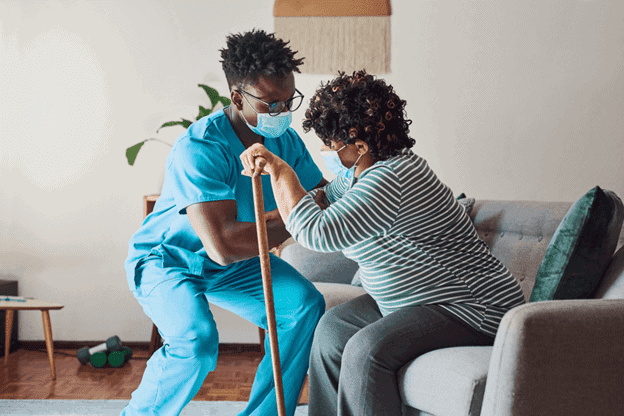 A home care aide helping and a senior woman stand, wearing masks