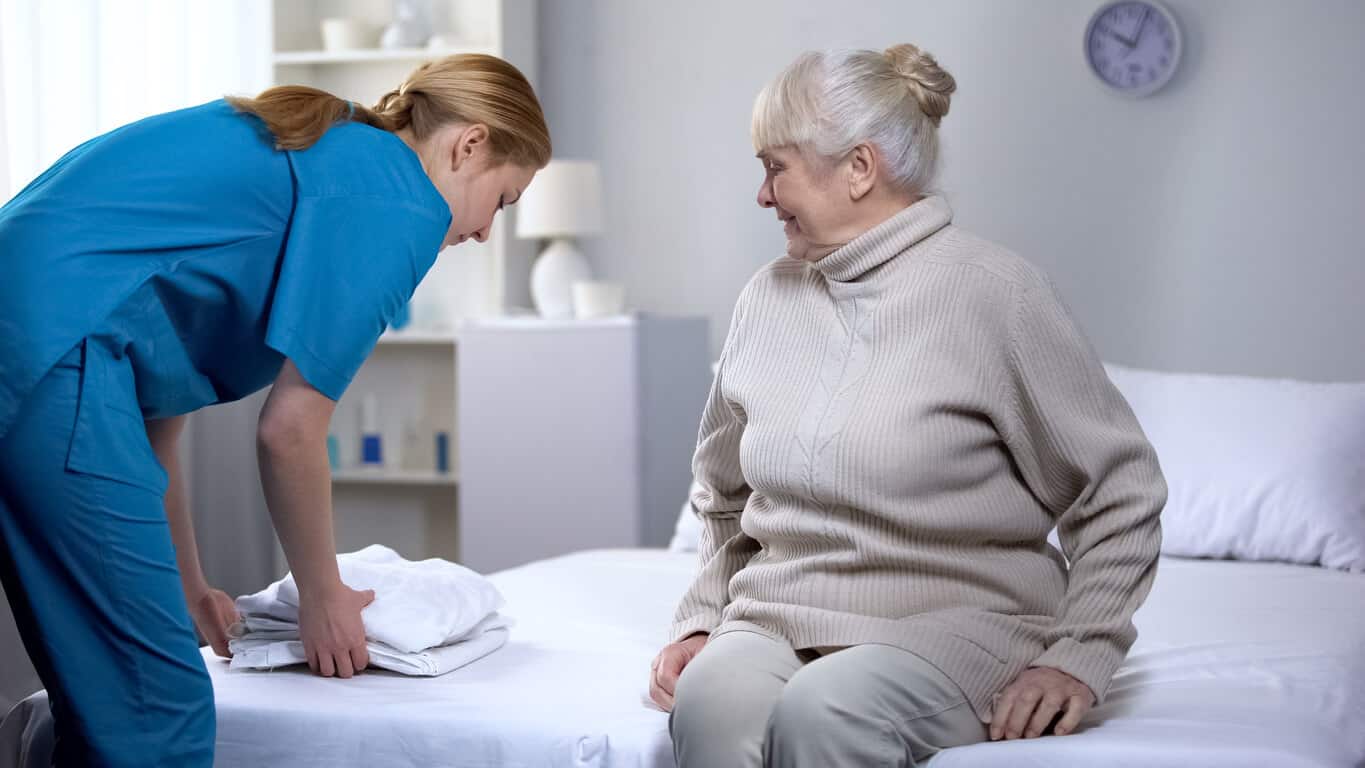 A caregiver providing home aid assistance by helping an elderly woman change her bed linens.