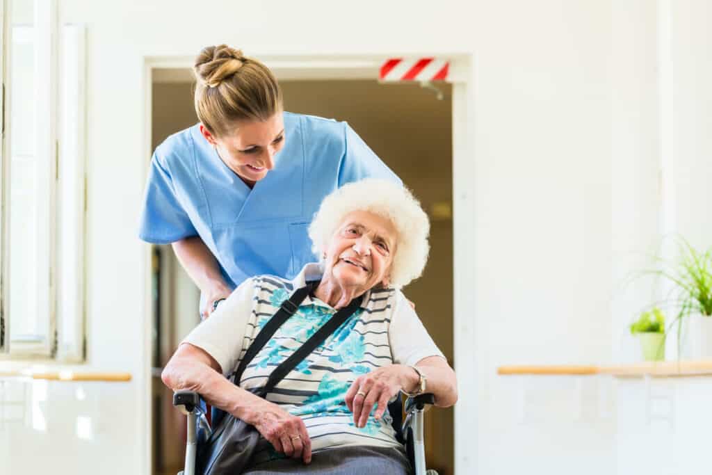 A smiling caregiver helping out a senior woman in a wheelchair after passing the caregiver certification course.