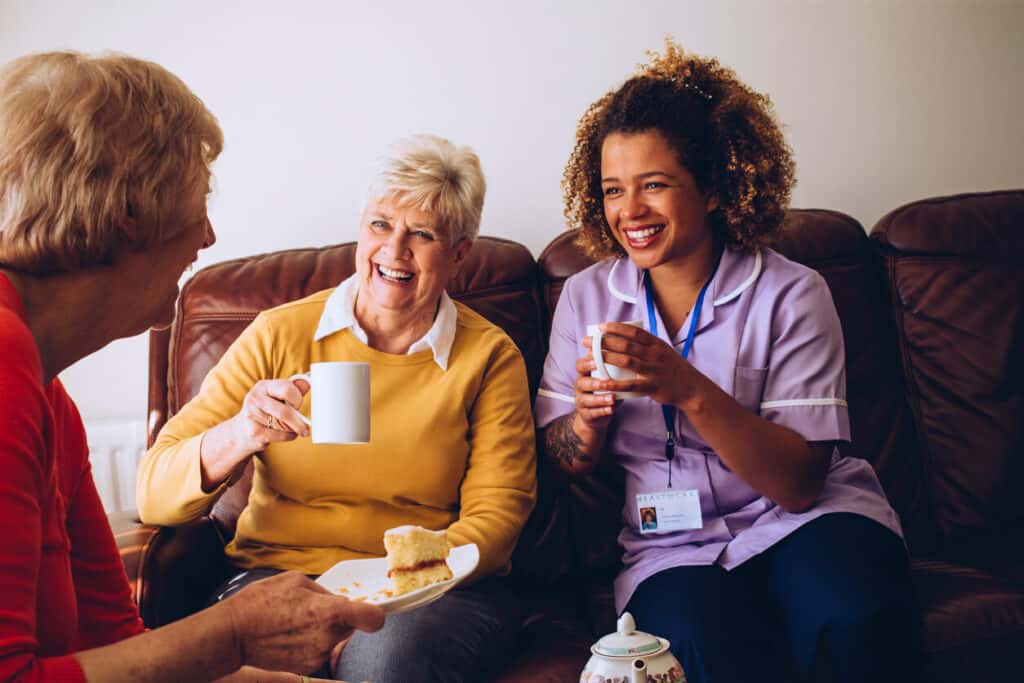 A certified caregiver sharing some cake and tea with two senior residents at a care home.