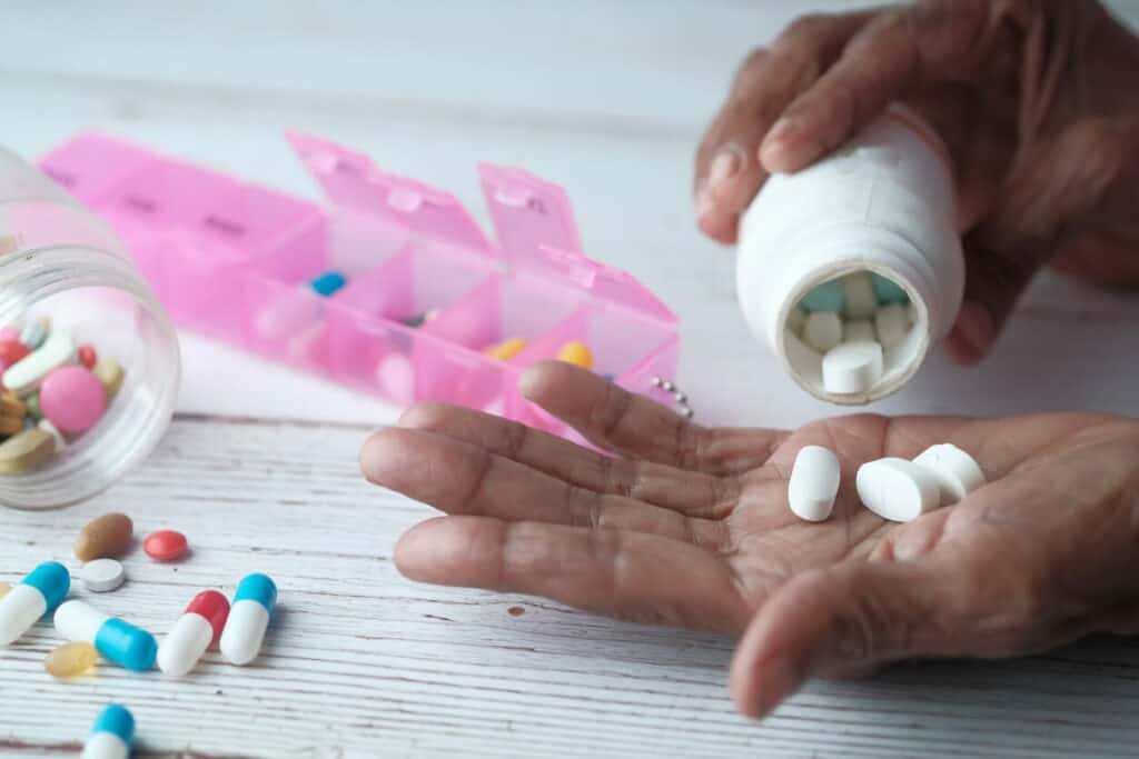 An elderly woman’s hand holding medicine pouring from a prescription bottle. Different colored pills lying on the table by her hand.