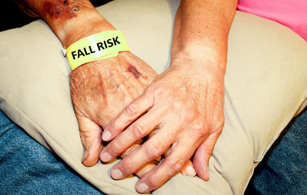 Two elderly hands, one laid over the other. One wrist has a yellow band that states “fall risk” on it.