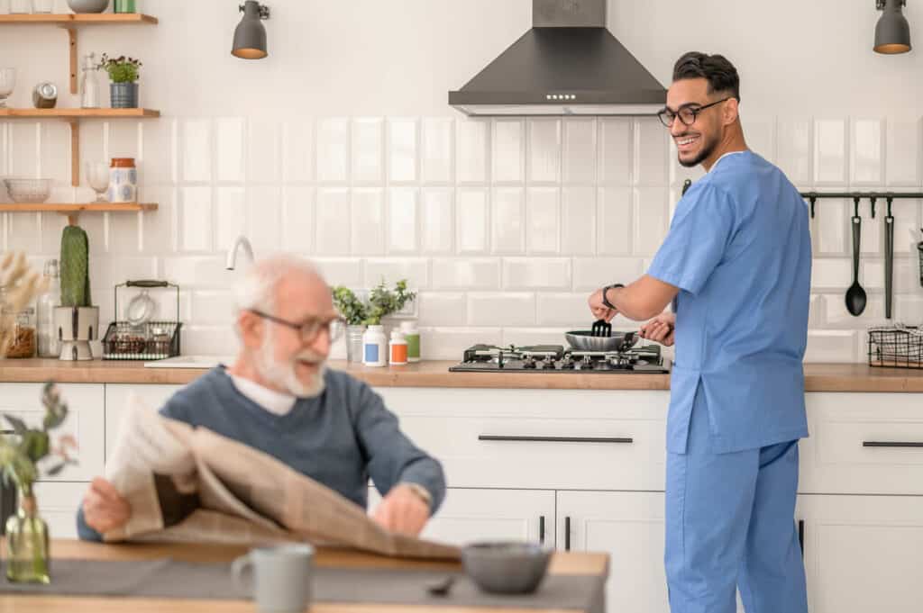 Male live-in caregiver preparing his senior’s breakfast while chatting with him