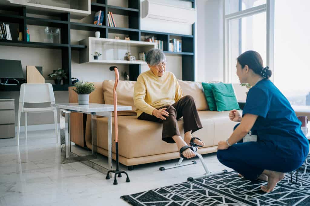 A geriatric occupational therapist teaching an older woman sitting on a couch some leg exercises.