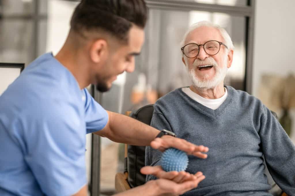 A male geriatric occupational therapist teaching a senior man hand exercises to regain hand strength.