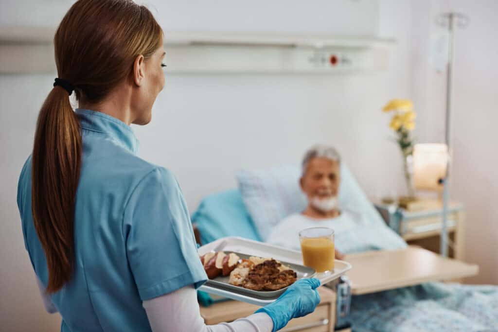 a smiling nurse taking food to a senior in bed using an overbed table