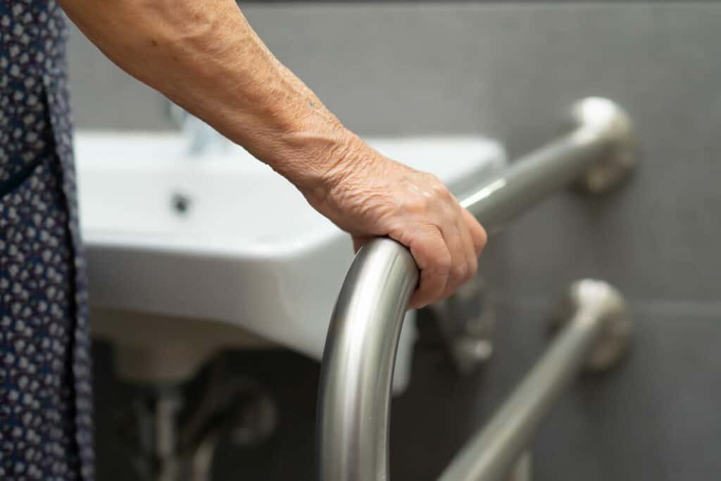 Senior lady grabbing a handrail while using her bathroom sink. Installing handrails is a good way to provide home care for Parkinson’s patients.
