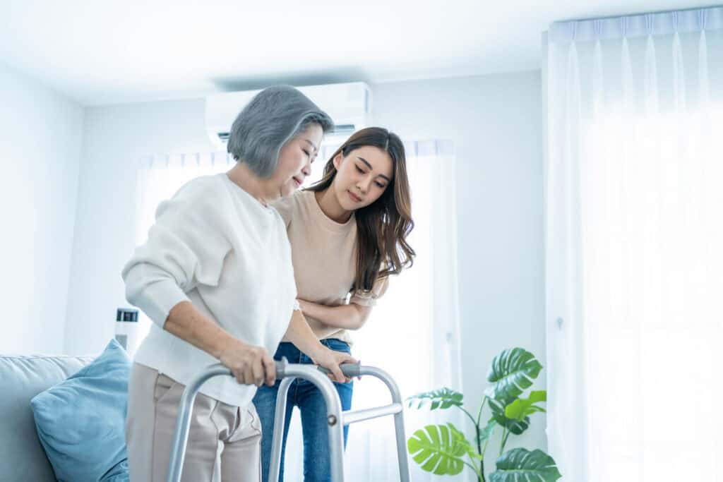 Asian grand-niece helping her senior aunt with Parkinson’s disease keep herself active by walking around the house. | Parkinson’s homecare