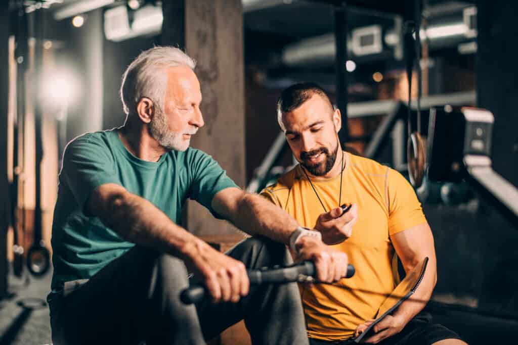 fitness instructor checking elderly gentleman’s heart rate during cardio exercise on a rower