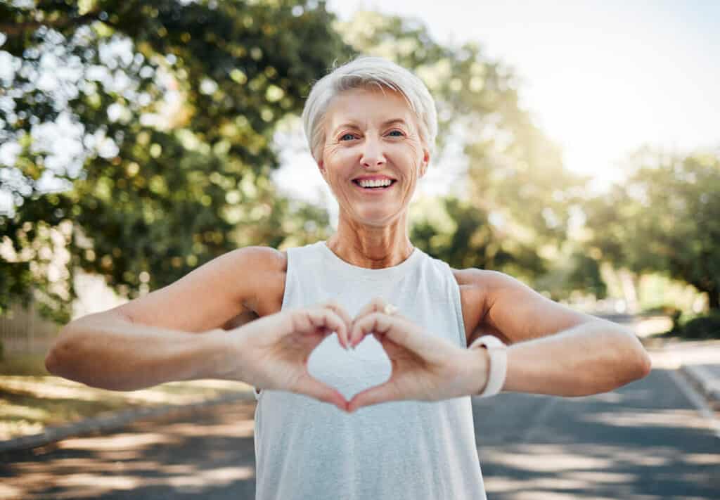 An elderly woman smiling and making a sign of heart after doing exercise in nature - aerobic exercises for seniors