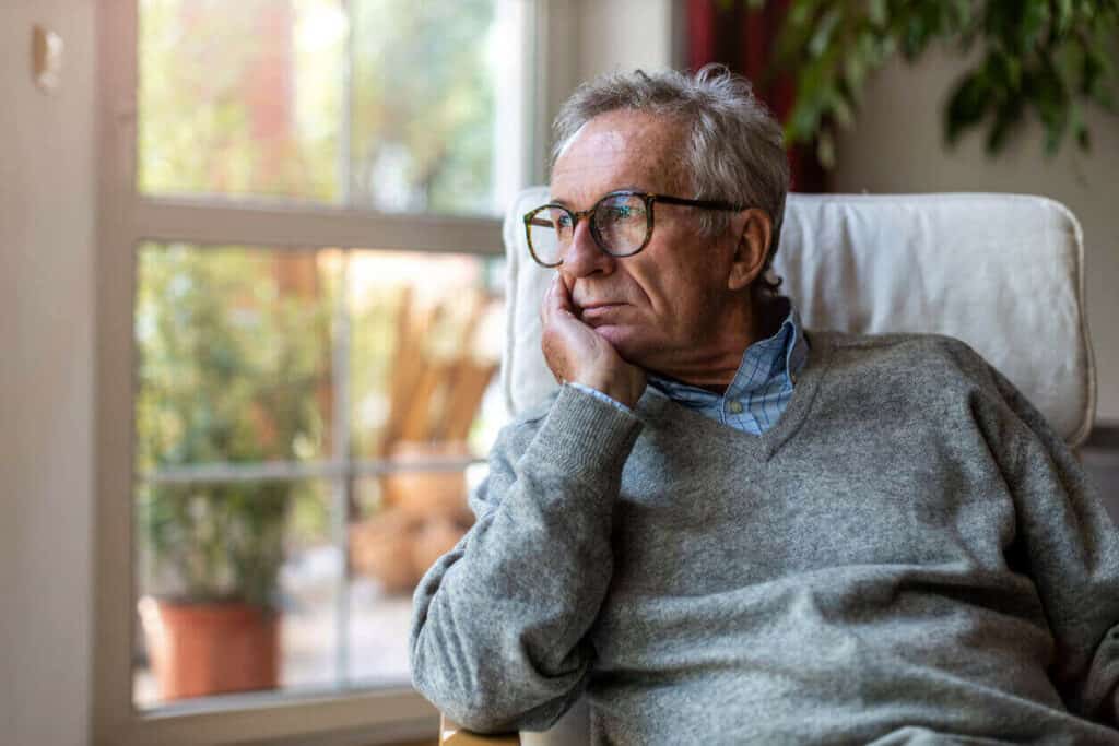 A senior man looking melancholically outside of his home. | A senior citizen hotline can help caregivers deal with elderly loneliness and depression.