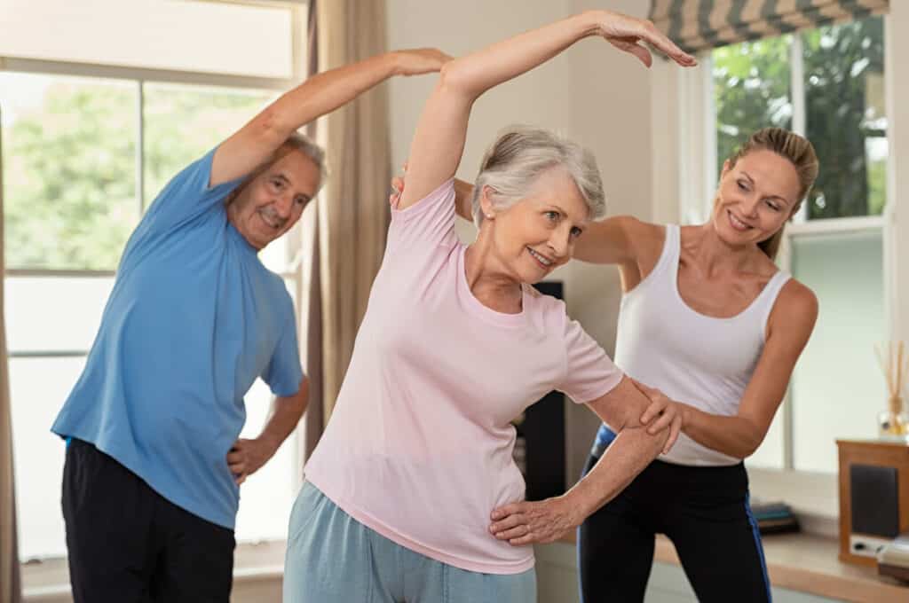 Elderly exercise benefits - happy senior couple doing exercise at home with their caregiver.