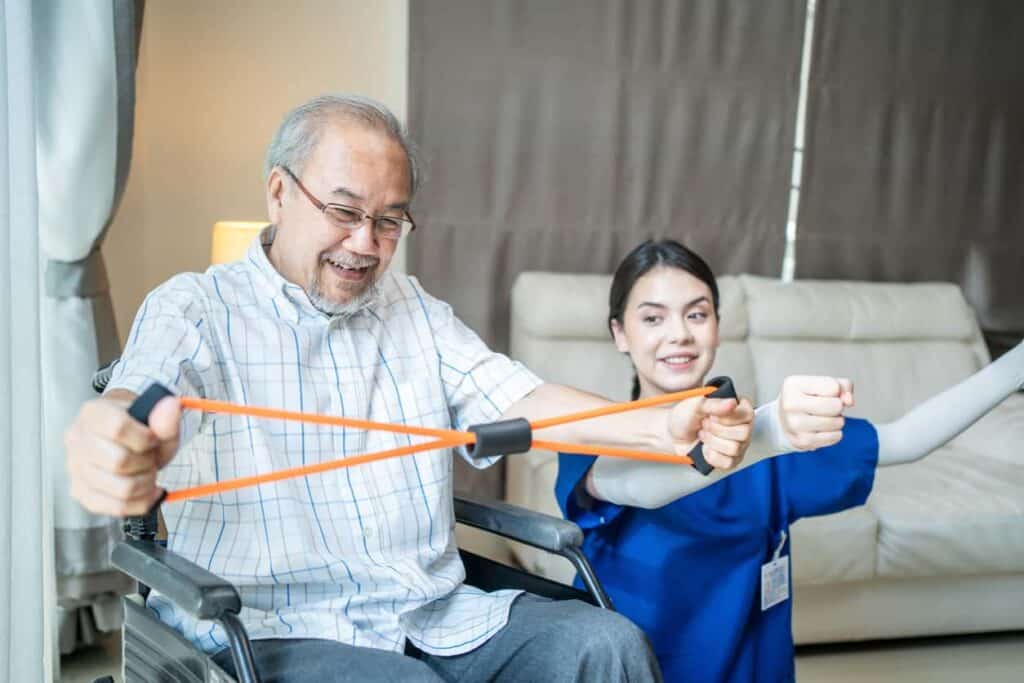 Disabled Asian senior man sitting on a wheelchair and stretching a rubber band while a female caregiver assists him in an assisted living facility