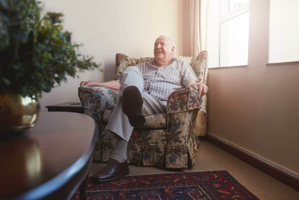 Elderly man laughing and sitting on his favorite couch that was brought to his nursing home room along with a plant that is being used as decoration for his table
