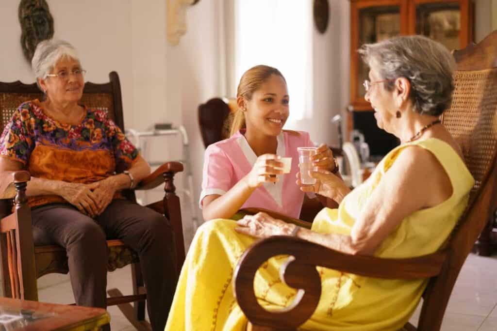 Latino woman caregiver giving water and medicine to an elderly woman while another senior female talks to them in a nursing home living room