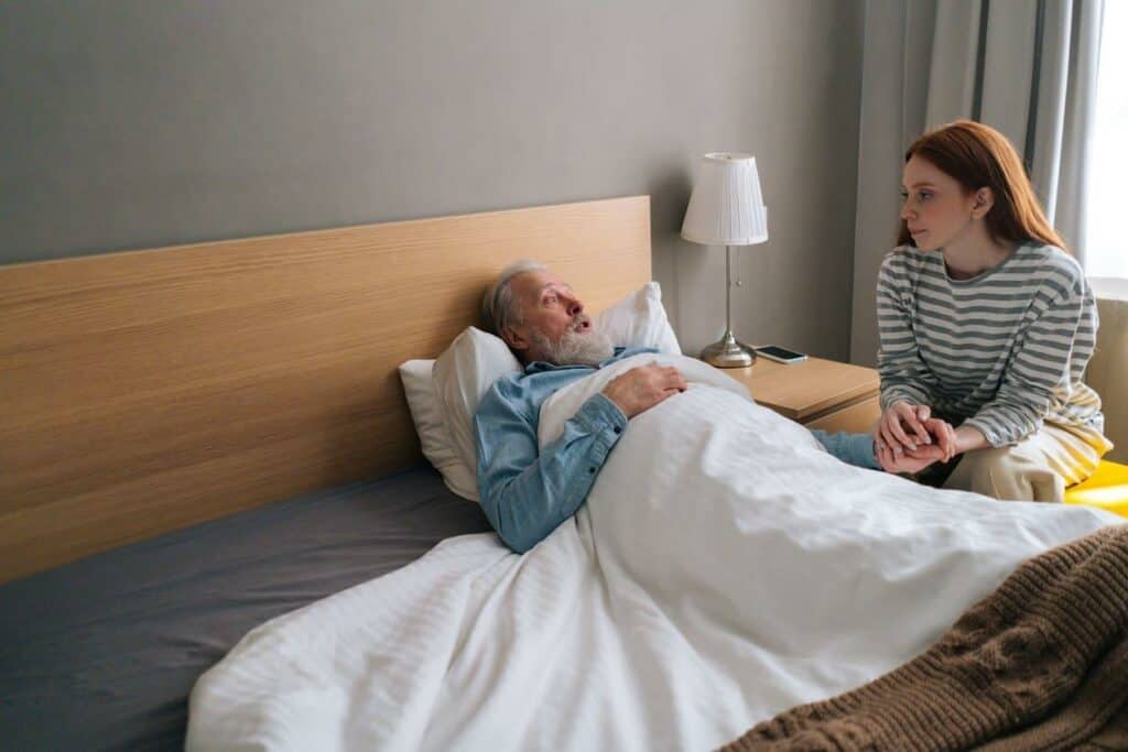 Female caregiver sitting by an older man in bed under palliative care - palliative treatment