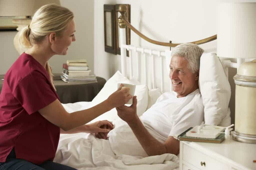 A caregiver handing a mug to a bedridden male senior patient - palliative care clinic.