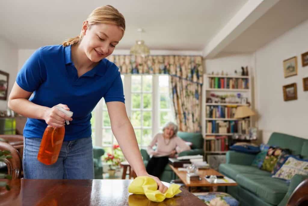 Home caregiver is cleaning the table while the elderly woman is sitting on the sofa in the background - homecare jobs