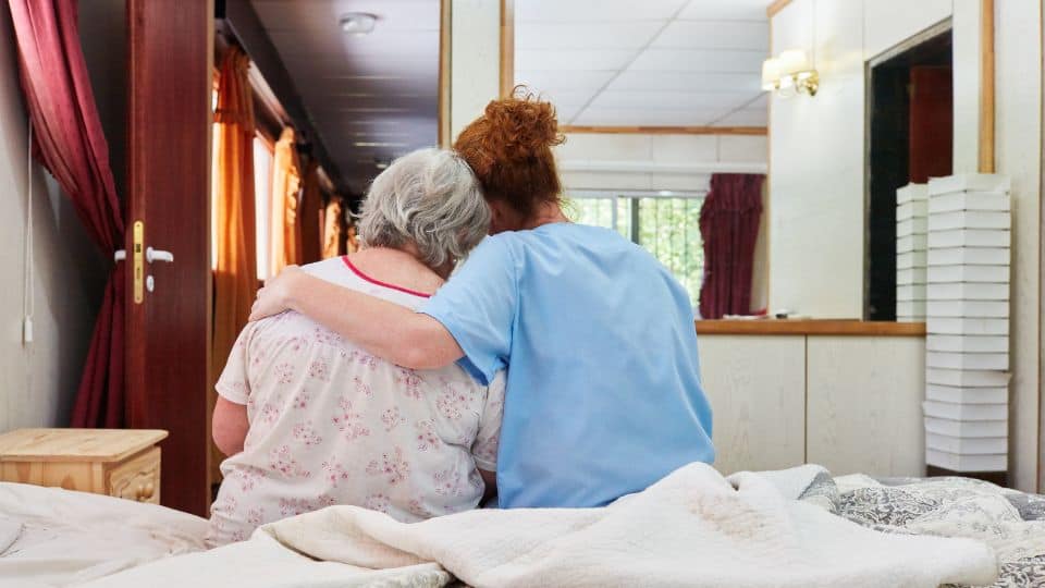 The back view of a nurse hugging a senior patient sitting on the bed.