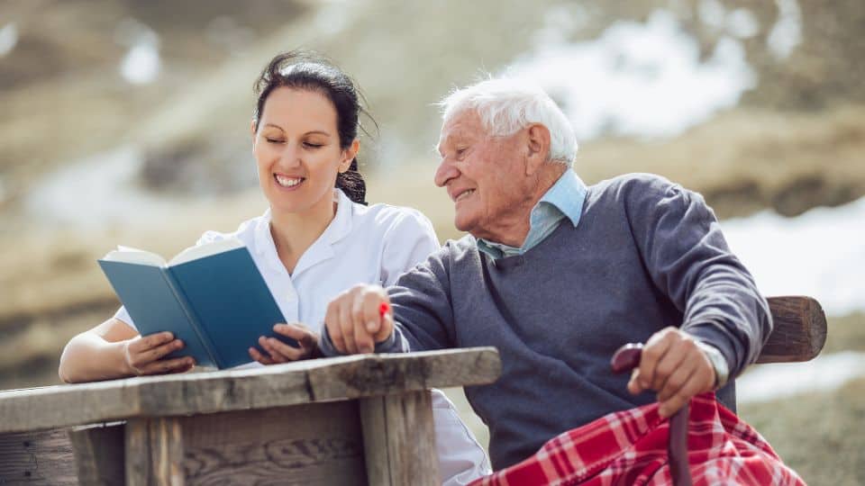 A caregiver reading a book to a senior patient outdoor.