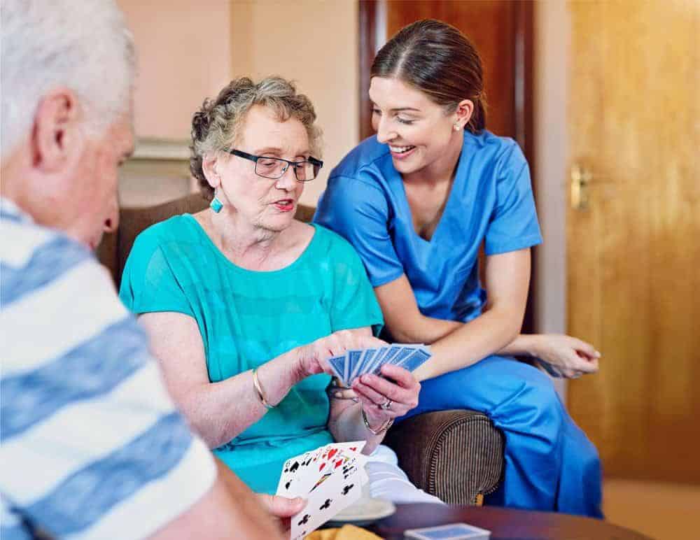 how to qualify for assisted living - senior man and senior woman with caregiver playing cards