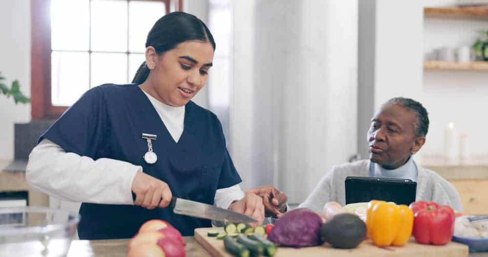 Female caregiver cooks a meal for an elderly man while listening to him talk | private in-home care