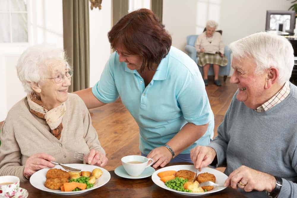 two seniors having a meal and laughing with their caregiver | independent assisted living