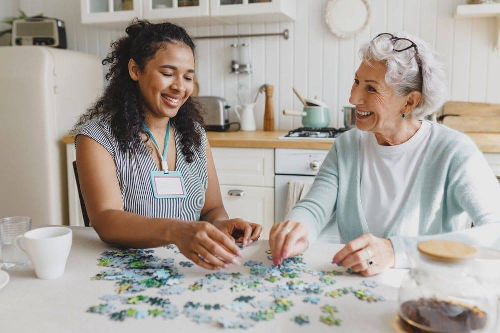 A senior woman accompanied by a 24/7 in home caregiver having fun and playing puzzles | how much does 24 hour inhome care cost