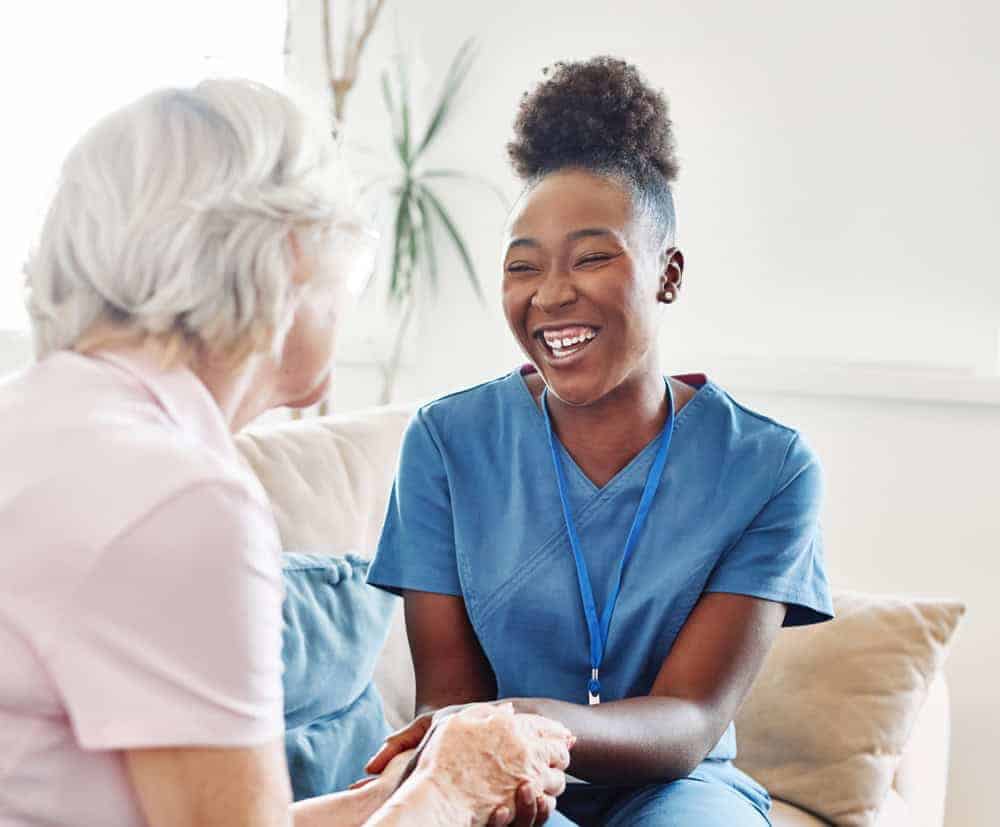 A woman nurse caregiver smiling while holding the hand of a senior woman - cost of live-in caregiver | cost of home health care